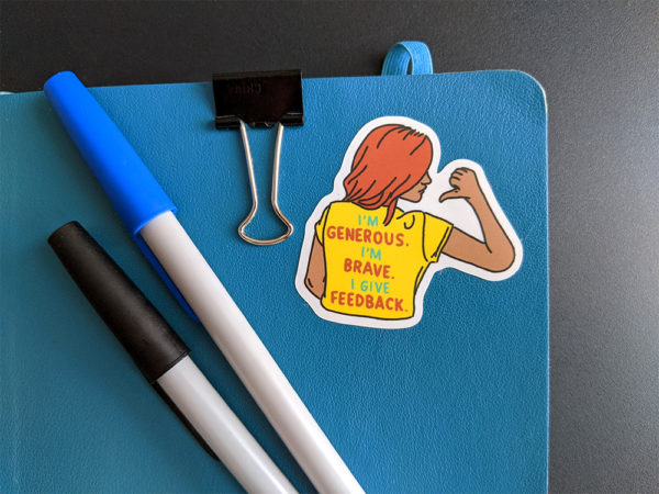 Sticker of woman wearing yellow shirt with "I'm generous, I'm brave, I give feedback" written on the back stuck to a blue notebook alongside two pens